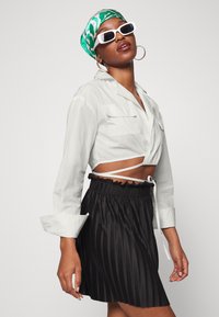 Camiseta branca cropped com dois bolsos no peito, combinada com uma saia preta plissada com um cós com volantes. O modelo usa um lenço verde e óculos de sol brancos.
