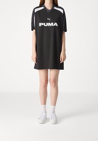 Czarna sportowa sukienka z krótkimi rękawami, z białym logo Pumy z przodu i szarymi akcentami na ramionach. Noszona z białymi sneakersami.