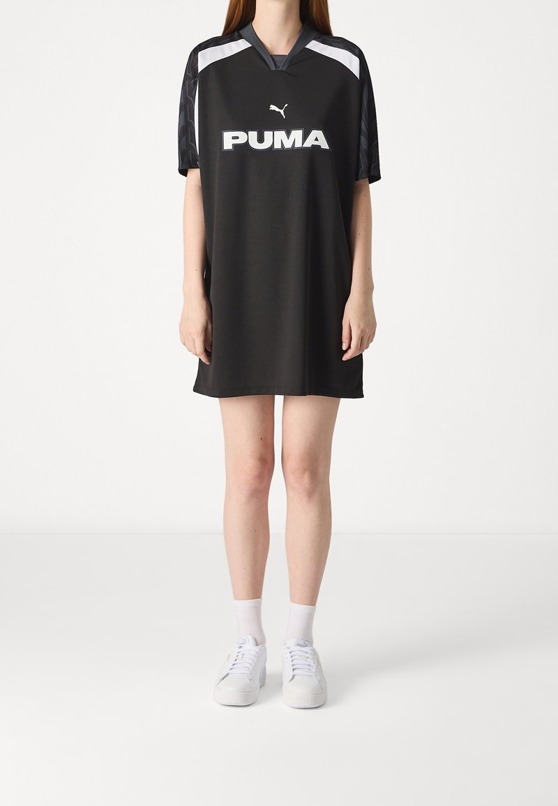Czarna sportowa sukienka z krótkimi rękawami, z białym logo Pumy z przodu i szarymi akcentami na ramionach. Noszona z białymi sneakersami.