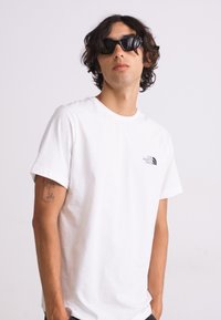 Hvid bomulds t-shirt med korte ærmer og et lille sort logo på brystet. Båret med sorte solbriller, stående mod en neutral baggrund.
