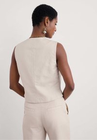 Beige ärmelloses Top aus Leinen mit taillierter Passform und geradem Rückennäht. Kombiniert mit passenden beigen Hosen mit Taschen auf der Rückseite.