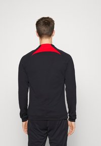 Camiseta deportiva de manga larga en negro con un acento rojo en los hombros. Fabricada con tejido elástico, presenta un diseño suave y ajustado.