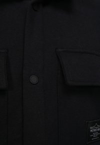 Zwarte gebreide shirt met knoopsluiting, twee voorkant zakken en een stoffen label met logo en tekst. Gladde textuur met een gestructureerd ontwerp.
