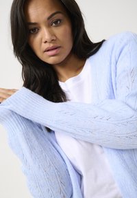Cardigan en tricot bleu clair avec une coupe ample, présentant des motifs texturés et des poignets côtelés, porté par-dessus une chemise blanche.