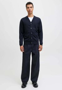 Uomo in piedi che indossa un cardigan nero con bottoni sopra una camicia bianca, pantaloni neri larghi e scarpe nere su uno sfondo chiaro e uniforme.