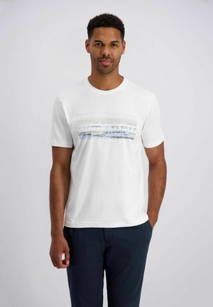 Mann mit kurzen lockigen Haaren, der ein weißes T-Shirt mit Grafik und dunkle Hosen trägt, steht vor einem schlichten hellen Hintergrund.