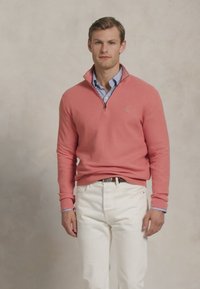 Jonge man in een koraalkleurige sweater met kwartrits over een blauw gestreept shirt, gecombineerd met witte broek, staande tegen een neutrale achtergrond.