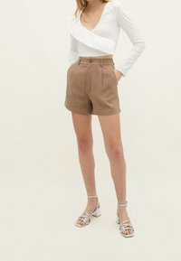 Lichtgewicht wit top met lange mouwen en een ruchtdetail, gecombineerd met khaki shorts met hoge taille. Model draagt zilveren sandalen met bandjes en blokhakken.