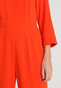Combinaison orange vif à manches longues avec une taille cintrée, montrée de la moitié du torse à la main sur un fond clair uni.