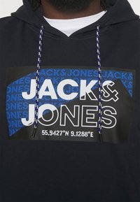 Marinblå hoodie med ett stort grafiskt tryck med "JACK & JONES" i vitt och blått, med koordinater längst ner. Flätade dragsnören ingår.