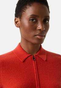 Roter gestrickter Pullover mit Reißverschluss und Kragen, strukturierter Stoff und schwarzer Reißverschluss-Akzent am Hals. Nahaufnahme des Kleidungsstücks.