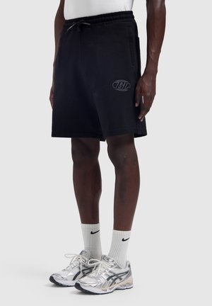 Person wearing black drawstring shorts with side pockets, white Nike socks, and silver running shoes, standing against plain background.