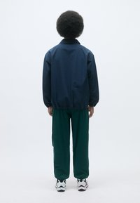 Marineblå vindjakke med en avslappet passform, elastiske mansjetter og rett snitt; kombinert med grønne bukser og hvite joggesko. Minimalistisk design.