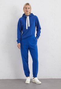 Blauwe hoodie met een witte koord en bijpassende blauwe sweatpants. Zachte stof, relaxed fit, ribbels aan de mouwen. Witte sneakers maken de look af.