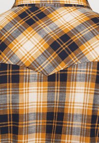 Primo piano di un tessuto con un motivo a quadri giallo, nero, bianco e marrone, che presenta un dettaglio a yoke appuntita sulla parte posteriore di una camicia.