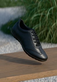 Pantof cu piele neagră, cu o finisare netedă, vârf rotund, design cu șireturi și accente texturate subtile pe laterale, așezat pe o suprafață de lemn.