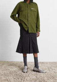 Camicia verde oliva con bottoni e due tasche sul petto, gonna nera a lunghezza ginocchio con orlo sfilacciato, mocassini grigi e calze grigie.