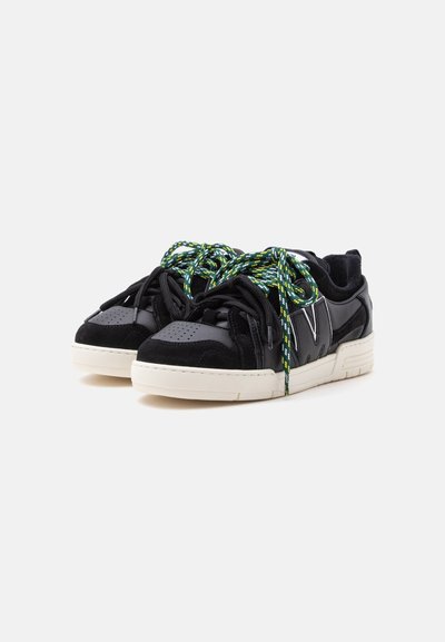 Ζευγάρι μαύρα low-top αθλητικά παπούτσια με λευκές σόλες και χοντρές κορδόνια με πράσινες, κίτρινες και μαύρες ρίγες σε λευκό φόντο.