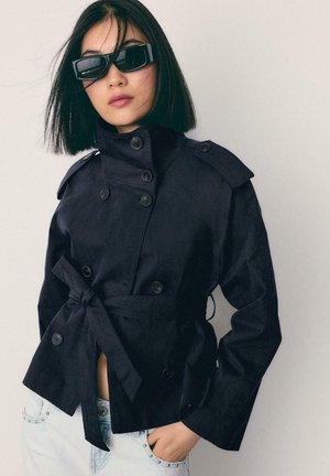 Frau mit kurzen schwarzen Haaren, die eine schwarze, doppelreihige Jacke mit Gürtel und schwarze rechteckige Sonnenbrille trägt, Hand in der Tasche einer hellblauen, mit Nieten verzierten Jeans.