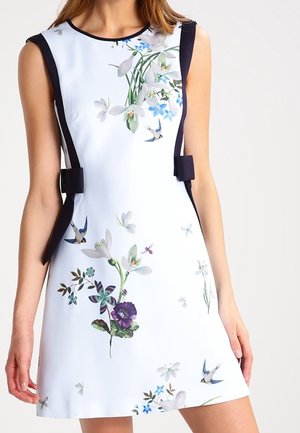 Sleeveless white dress with black side panels, decorated with colorful floral and bird prints, worn by a standing woman.