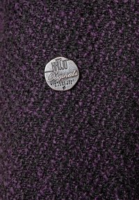 Tela texturizada en púrpura y negro con una etiqueta metálica redonda que dice "KHUJO Originals inspirados en la moda vintage de NYC."