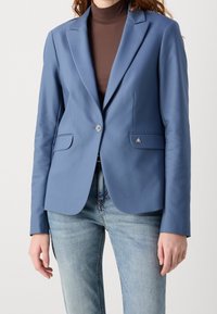 Žena v modrém na míru šitém saku přes hnědý rolák a světle modré džíny, stojící na jednobarevném pozadí.