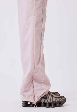 Lichtroze broek met verstelbare trekkoorden bij de boorden, gecombineerd met grijze en donkerbruine sportschoenen met veerachtige zolen.