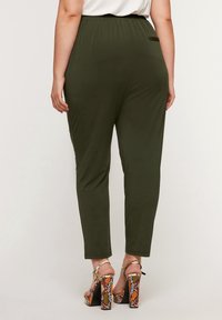 Nainen, jolla on yllä oliivinvihreät kapeat housut ja korkokengät, joissa on oranssin- ja ruskeankirjava käärmeennahkakuosi, seisoo yksinkertaista taustaa vasten.