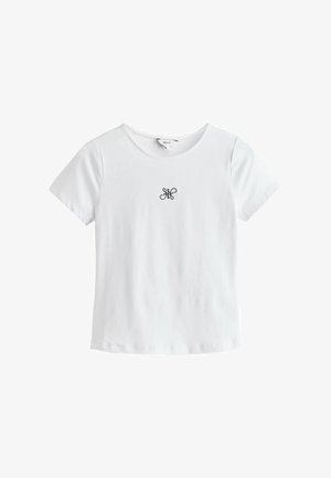 Fehér rövid ujjú póló kerek nyakkivágással és kis fekete díszítő emblémával a mellkas közepén.