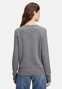 Frau mit langen braunen Haaren, die einen grauen Langarmpullover und bluejeans trägt, von hinten vor einem schlichten Hintergrund gezeigt.