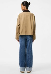 Rjava srajčna jakna s temnim ovratnikom, kombinirana s širokimi modrimi denim hlačami z rabo robom in svetlo modrimi manšetami. Minimalistična zasnova.