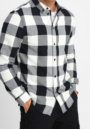 Homme portant une chemise à carreaux noire, blanche et grise avec les mains dans les poches d'un pantalon noir, cadré au niveau du cou.