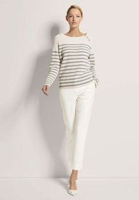 Gestreifter Strickpullover in Creme und Grau, mit einem weiten Rundhalsausschnitt und Knöpfen an einer Schulter. Kombiniert mit weißen, schmal zulaufenden Hosen.