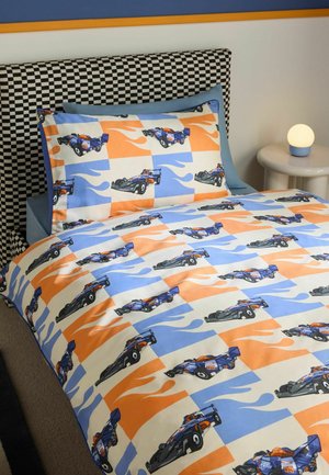 Einzelbett mit orange-blauer Bettwäsche mit Rennwagenmuster, kariertem Kopfteil, blauer Wand und rundem Nachttisch mit leuchtender Lampe.