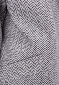 Hahnentrittgewebestoff in Grau und Weiß mit weicher Textur; zeichnet sich durch markante Zickzacklinien und ein gewebtes Aussehen aus.