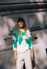 Weißer Pullover mit grünem abstraktem Muster, Rundhalsausschnitt und langen Ärmeln; beigefarbene Hose; strukturierter Steinwand-Hintergrund.