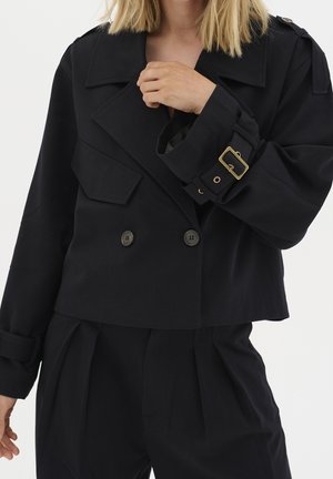 Femme portant une veste courte noire à double boutonnage avec poche à rabat et détail de boucle en or sur la manche.