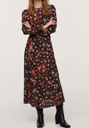 Kvinde iført en lang sort blomsterkjole med orange og røde blomster, kombineret med sorte ankelstøvler med hæl, armene krydsede.