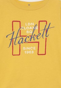 Žluté bavlněné tričko s modrým a červeným grafickým textem: "Hackett," "LDN SLOANE SQ," a "SINCE 1983" v různých písmech a velikostech.