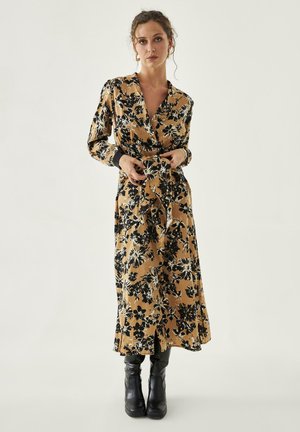 Mujer ajustando el cinturón de un vestido largo color beige con estampado floral negro, llevando botas negras, de pie contra un fondo claro y sencillo.