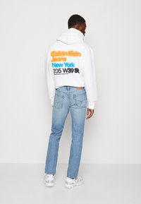 Hvit hettegenser med hette som har flerfarget tekst på ryggen, kombinert med lyse blå denimjeans og hvite joggesko.