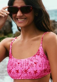 Rozā smockēts bikini tops ar plānām siksnām un ziedu rakstu. Struktūras materiāls rada sakrālu efektu, kombinējot ar sarkanām saules brillēm.