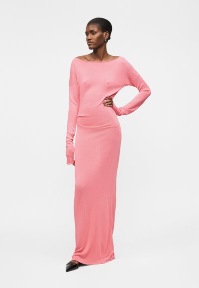 Γυναίκα στέκεται με το χέρι στη μέση, φοράει μακρυμάνικο ροζ φόρεμα μέχρι το πάτωμα και μαύρα μυτερά παπούτσια σε απλό λευκό φόντο.