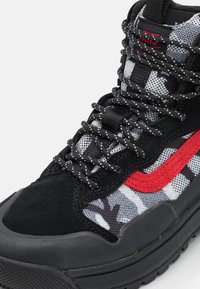 Baskets montantes en daim noir et mesh avec des accents rouges, textile à motifs, lacets noirs et semelle en caoutchouc avec une finition texturée.