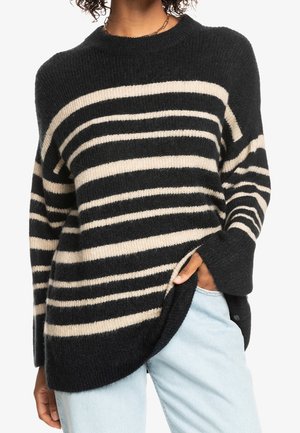 Pull à manches longues rayé noir et beige, porté avec un jean bleu clair, main dans la poche, présentant un col rond et une coupe décontractée.