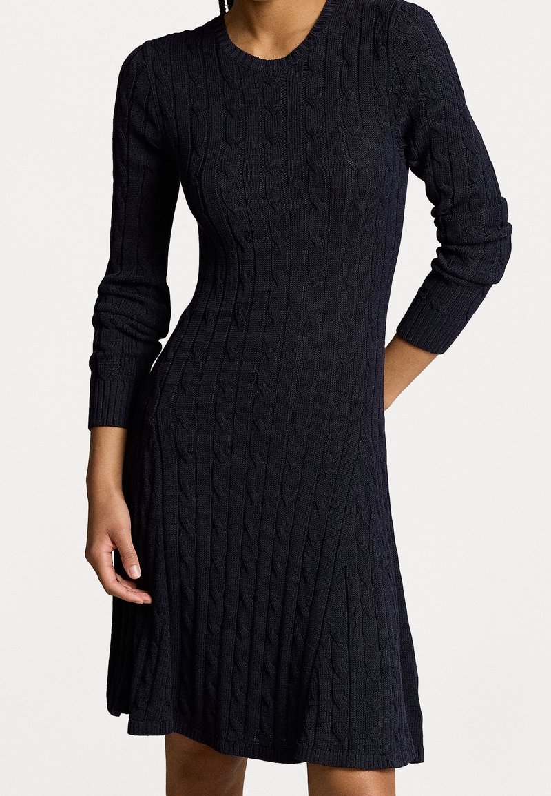 Femme portant une robe noire moulante en maille torsadée à manches longues avec un col rond, debout avec une main sur la hanche et une main détendue.