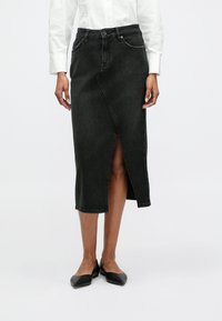 Jupe midi en denim noir avec une fente à l'avant, taille haute et passants de ceinture. Portée avec des chaussures plates noires et une chemise blanche à boutons.