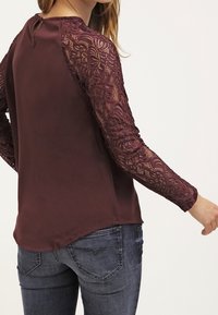 Burgundy pusero, jossa on pitkät pitsihihat, joissa on kukkakuviota. Sileäkangasrunko, jossa on hienovaraan kiiltoa, takana avainauko suljenta ja pyöristetty helma.