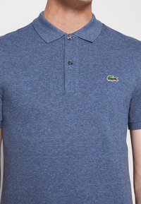 Blå pikétröja i strukturerad tyg, med spetsig krage, två knappar och en liten grön krokodil-logotyp på vänster bröst.