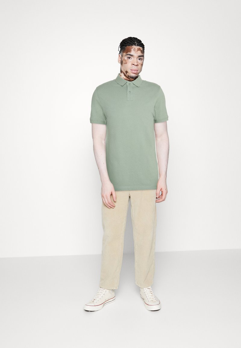 Zelená polo tričko s krátkým rukávem, kombinované se světle hnědými kordovými kalhotami a bílými teniskami. Jednoduchý design, měkká tkanina, volný střih.
